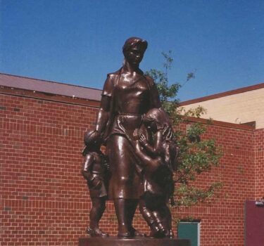 bronze sculpture of woman and two children atop cube granite base in front of brick building