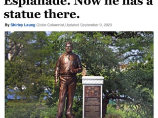 image of news article title and photo featuring statue of man standing on granite base surrounded by landscaping with river behind
