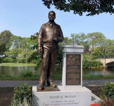 bronze statue of man beside podium with bronze plaque set in a park with lake behind