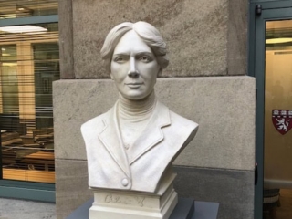 detail of white plaster bust of female, namely Alice Hamilton, on a gray pedestal in front of a stone wall between windows