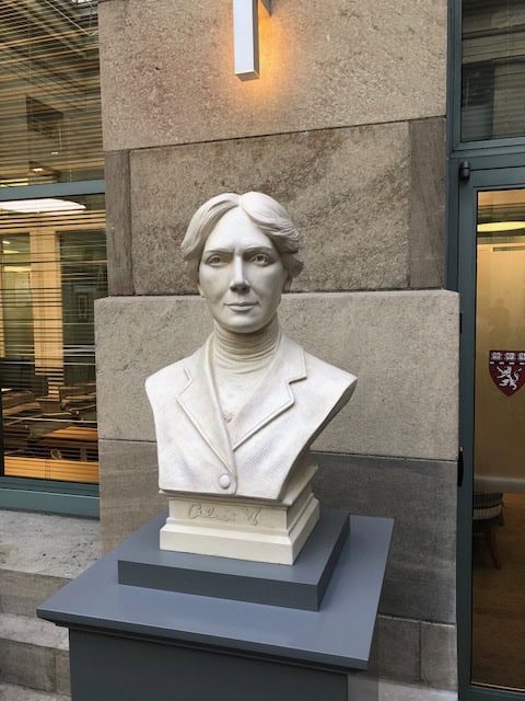 detail of white plaster bust of female, namely Alice Hamilton, on a gray pedestal in front of a stone wall between windows