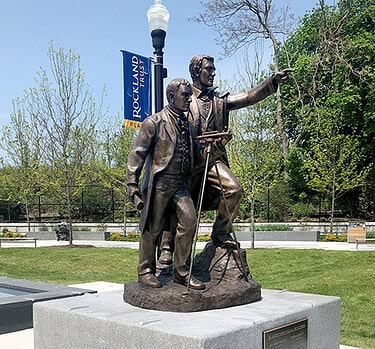 sculpture of two figures on square granite base in plaza with hardscaping and landscaping
