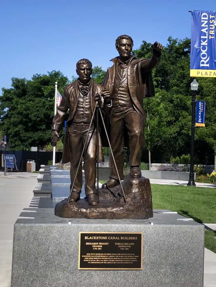 sculpture of two figures on square granite base in plaza with hardscaping and landscaping